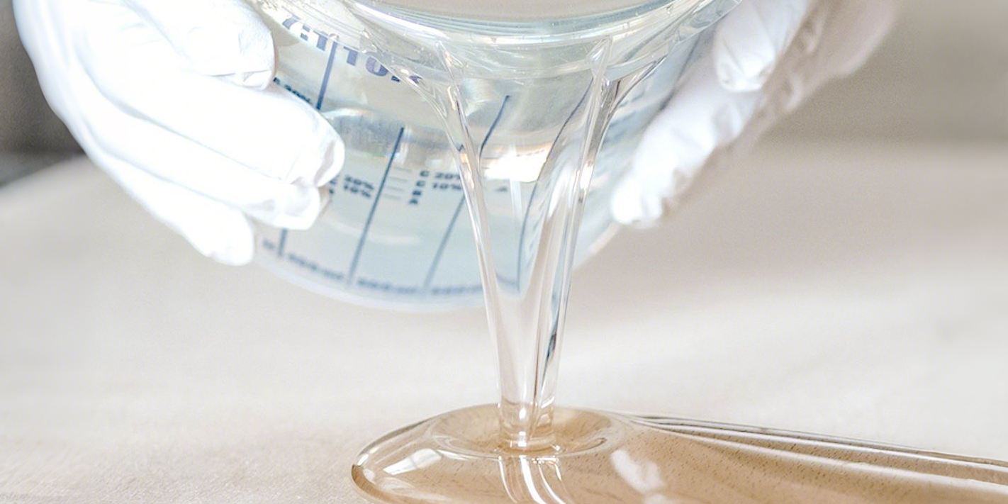 Gloved hands pour clear epoxy resin — a key material for deep pour epoxy resin projects — from a measuring cup, showcasing its glossy, viscous flow as it cascades onto the surface.