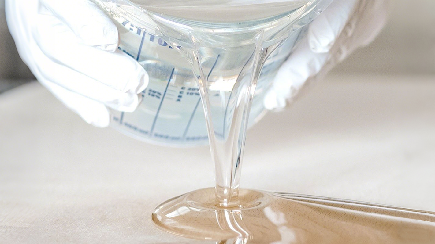 Gloved hands pour clear epoxy resin — a key material for deep pour epoxy resin projects — from a measuring cup, showcasing its glossy, viscous flow as it cascades onto the surface.